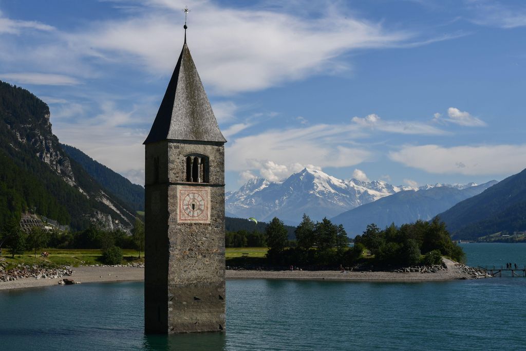 A picturesque view of a church steeple, old brick with a pitched roof, halfway submerged in water with a view of distant mountains and blue sky, wispy white clouds behind