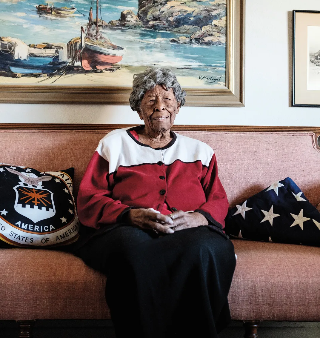 A Black woman sits on a couch surrounded by pillows and art on the wall in her home.