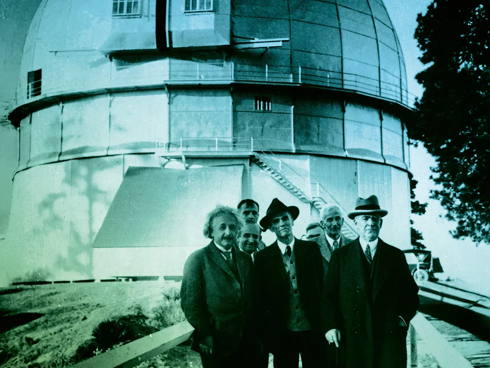 A black-and-white historical photograph from circa 1931 shows Albert Einstein with Edwin Hubble and other astronomers standing on a walkway in front of the large dome of the Mount Wilson Observatory.