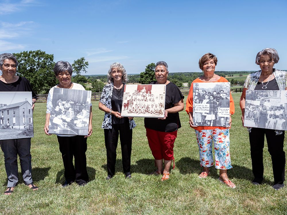 Six elder Black women pose on a grassy hillside, each holding an enlarged black-and-white photo of a schoolhouse, protestors with signs, and a class photo.
