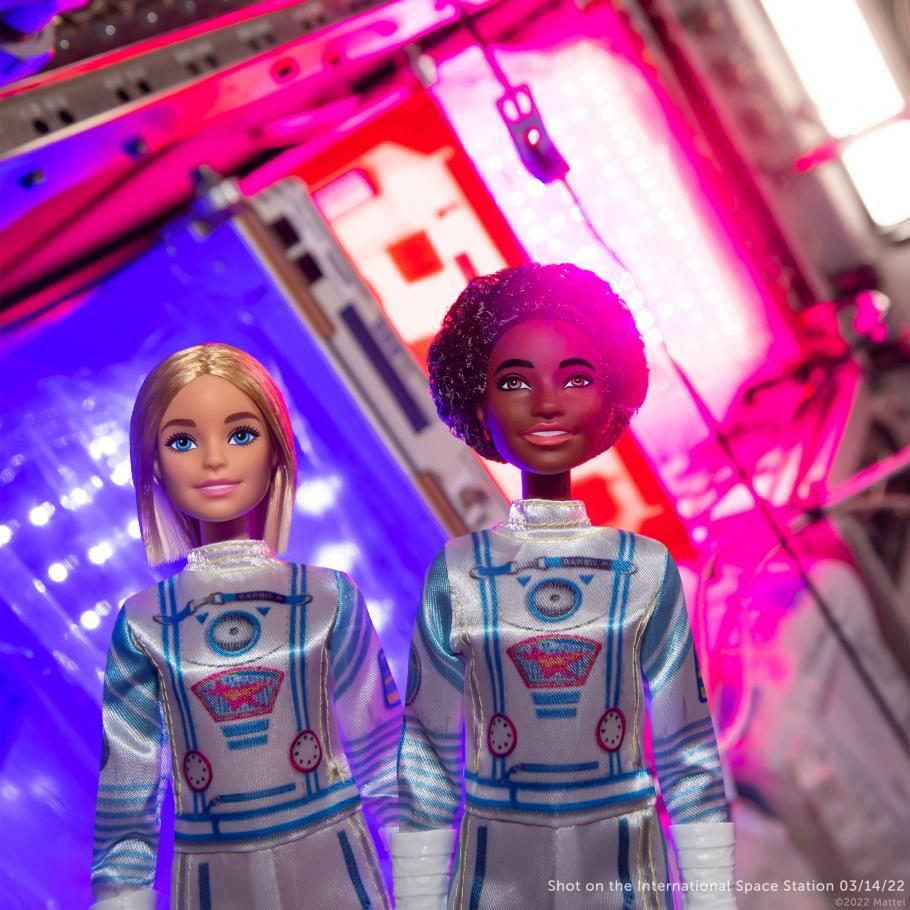 Two Space Discovery Astronaut Barbies photographed in front of the Veggie Garden on the International Space Station. Bright purple and pink lights can be seen behind them.