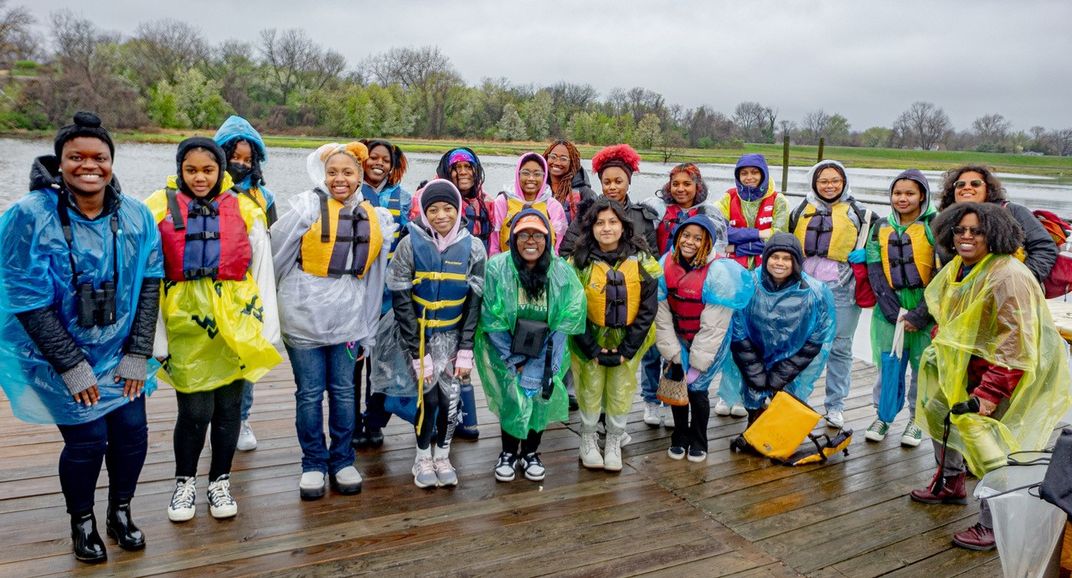 A group of young people of color pose on a dock next to a waterway, outfitted with life vests and waterproof ponchos.