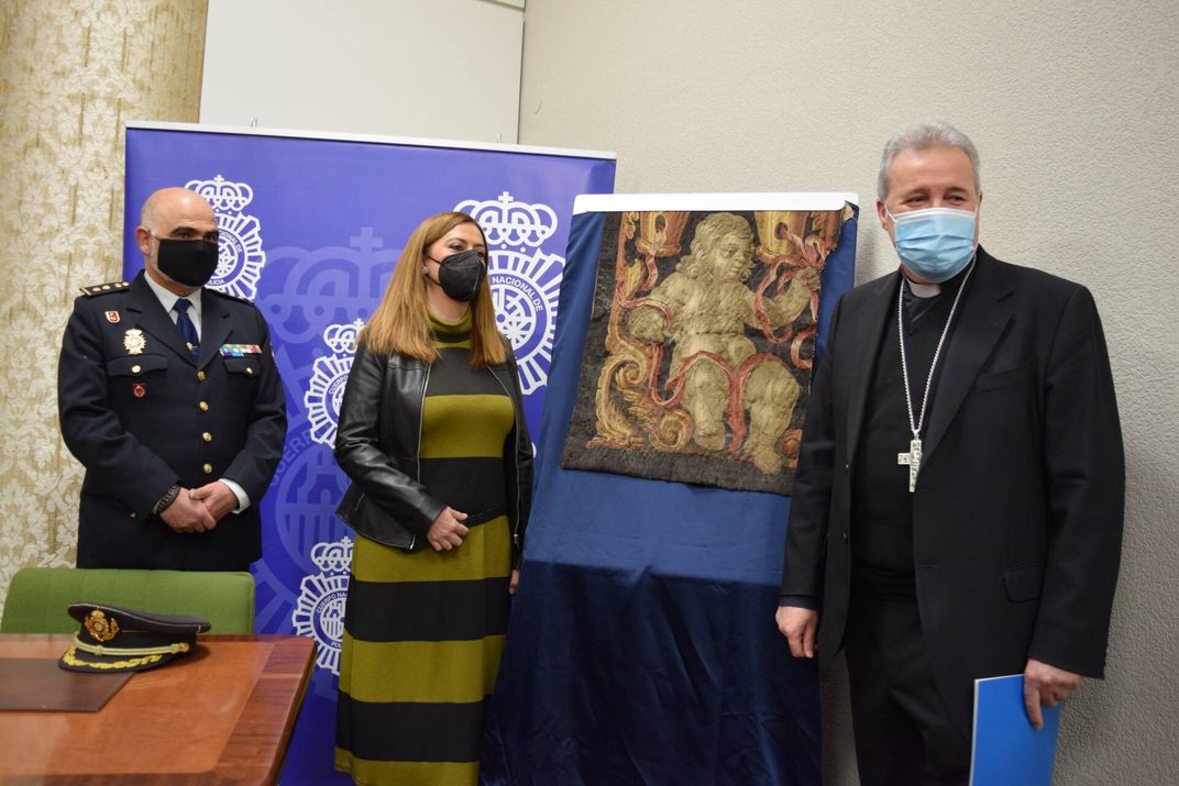 Three people in masks, including one priest and one police officer, stand near the square of tapestry at a formal press conference