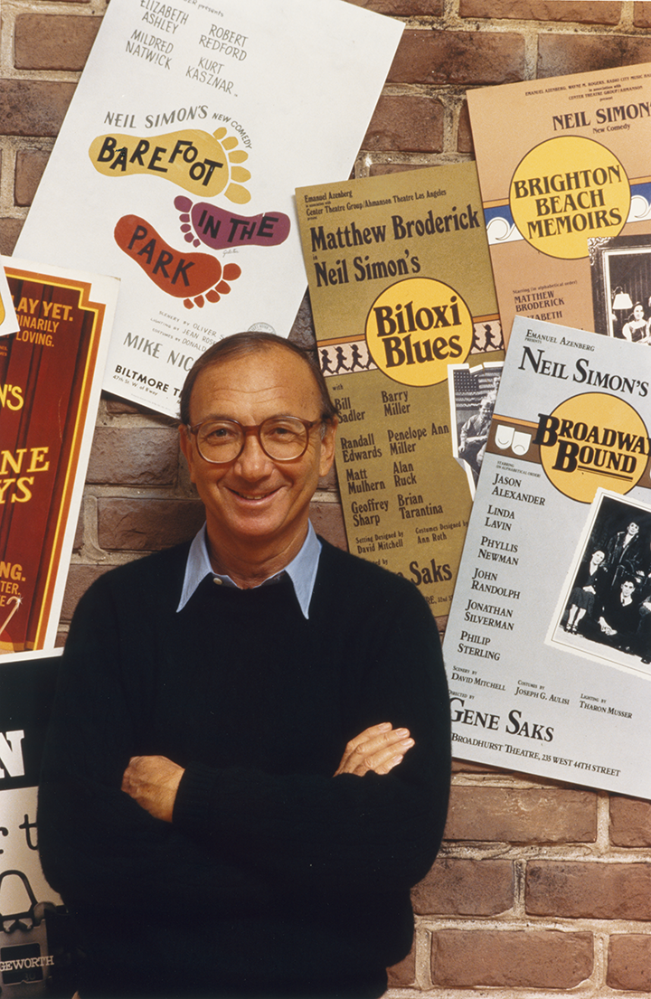 Neil Simon stands with arms crossed, smiling against a brick wall covered in posters