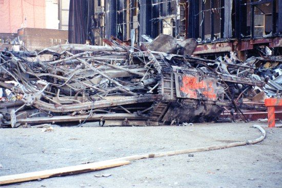 Photo of destruction in New York City after September 11, 2001. Collapsed industrial-looking part of a building with many pipes, columns, and wires. In background, broken windows. Ash and gray.