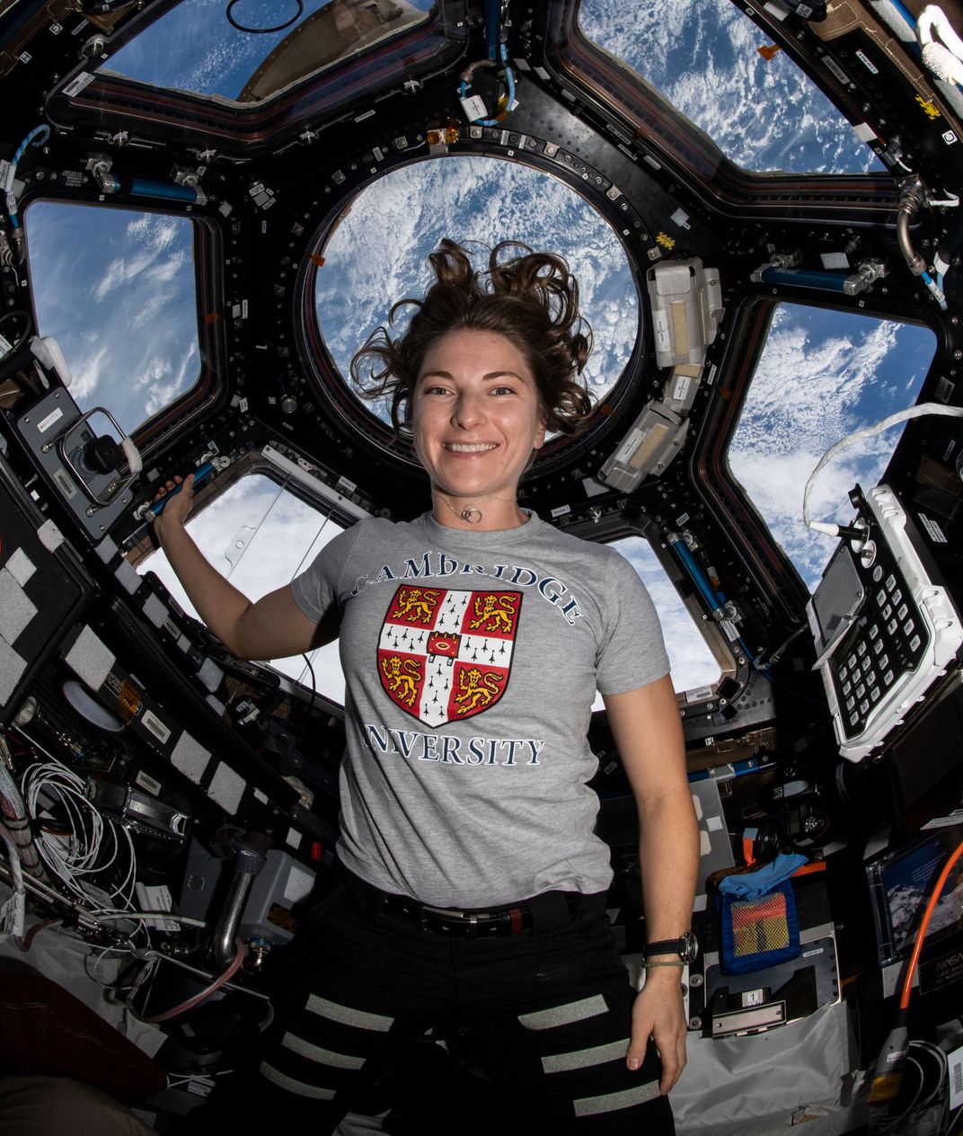 female astronaut posting next to a window at the space station, the earth pictured behind her