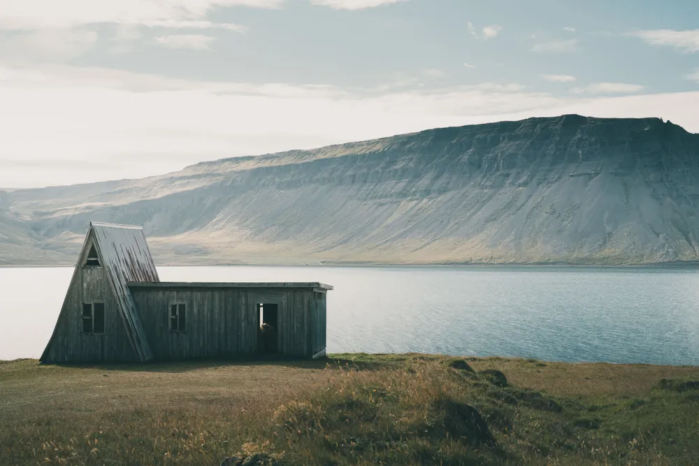 This rustic A-frame cabin stands quietly on the edge of a serene fjord, framed by majestic cliffs in the background. Its weathered wood tells a story of resilience against the elements in this remote, peaceful landscape.