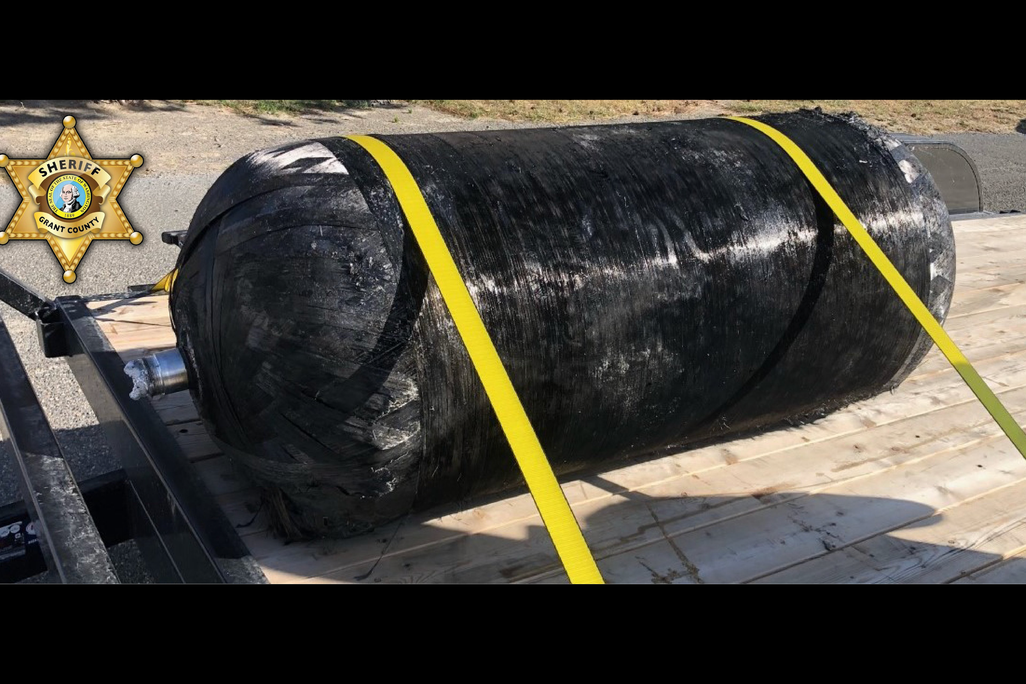 A photo of a large black cylinder strapped to a wood platform with yellow cords