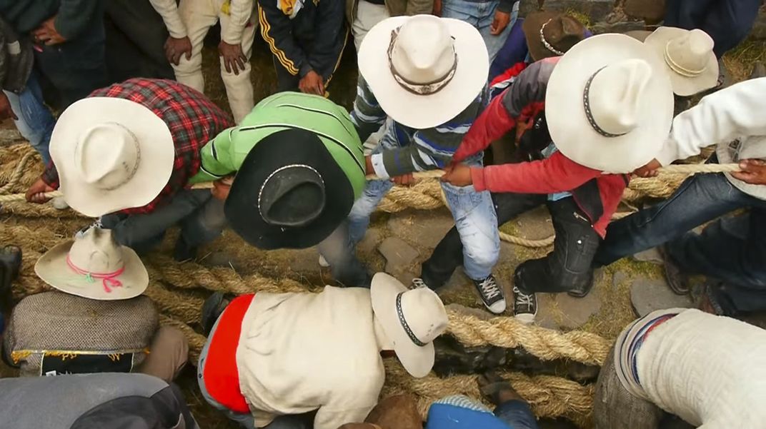 Men bend over to pull very large woven-grass cables taut. The perspective is from above, so what is seen is the crowns of the men's cowboy hats, their grip on the thick cables, and their position back on their heels as they pull.