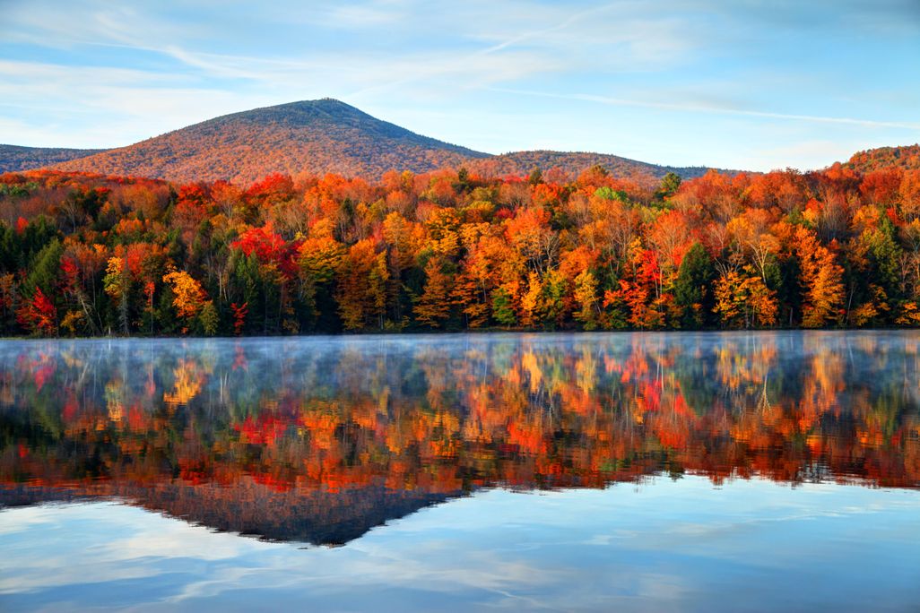 A lake, with a distant sloping hill and brilliant and orange red trees reflected perfectly in the still water. Overhead, a blue sky with wispy white clouds