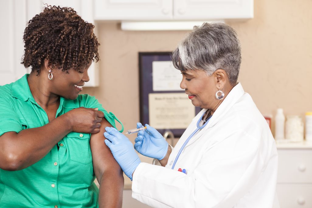 An image of a woman of color recieving a vaccine from a doctor. The doctor is a woman of color dressed in a white lab coat.