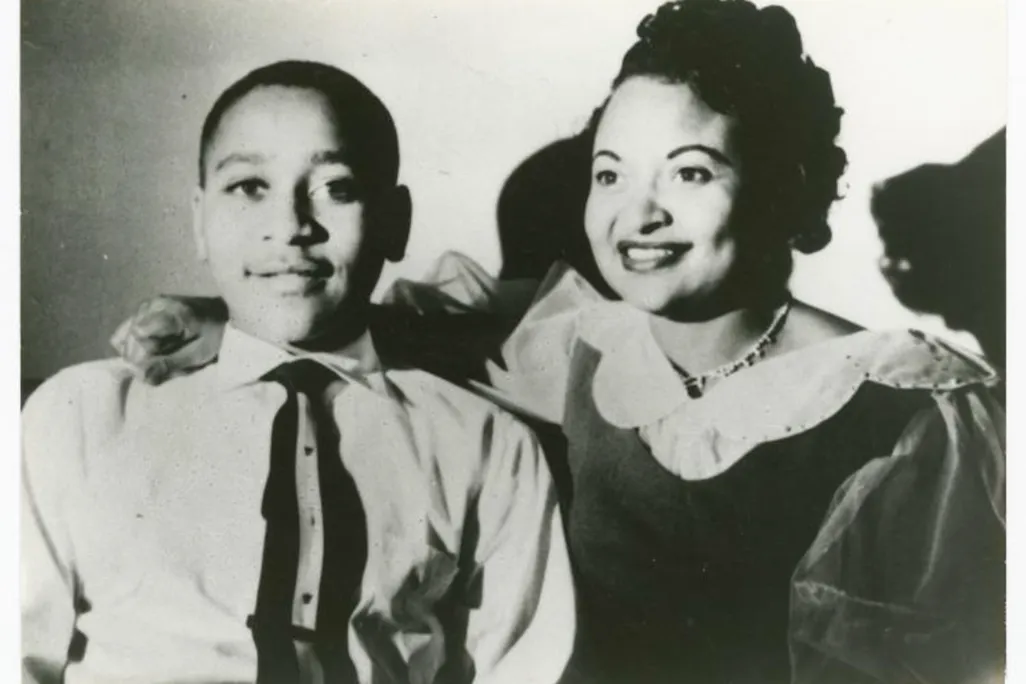 Emmett Till, a young boy in a suit and tie, smiles next to his mother, who wears a dress and has her hand on his shoulder