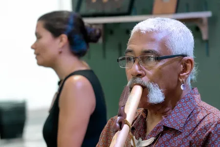 A man with short white hair, moustache, and glasses plays a wooden flute. Behind him, slightly out of focus, is a young woman with long dark hair in a bun and a black tank top.