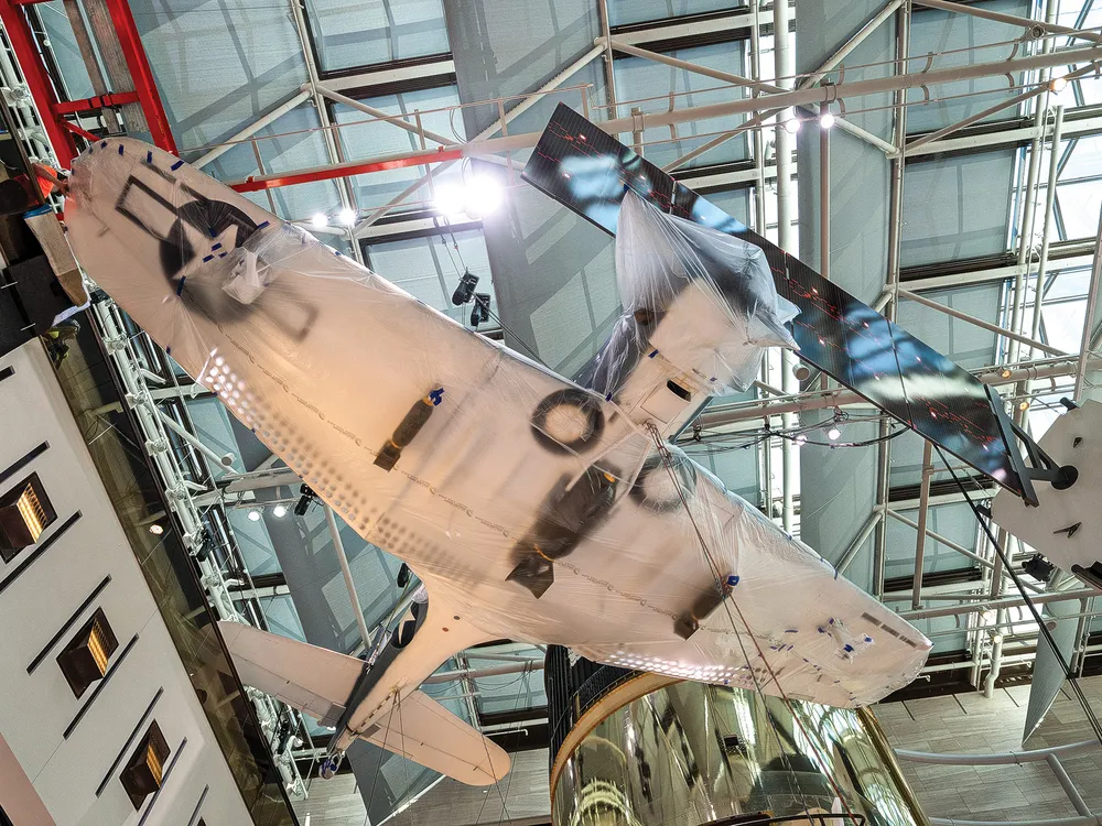 A World War II military airplane is photographed from below while hanging from the ceiling. The underside of the airplane is painted white and the wing and forward fuselage are wrapped in opaque plastic.