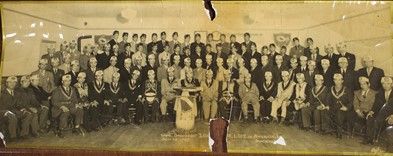 50 plus men in corresponding lodge regalia are posed in four staggering rows in a gathering hall.