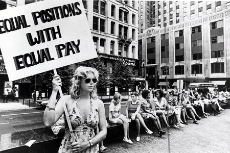 Woman holding a sign reading “Equal positions with equal pay” during an equal pay demonstration in a city plaza, with women seated behind her.