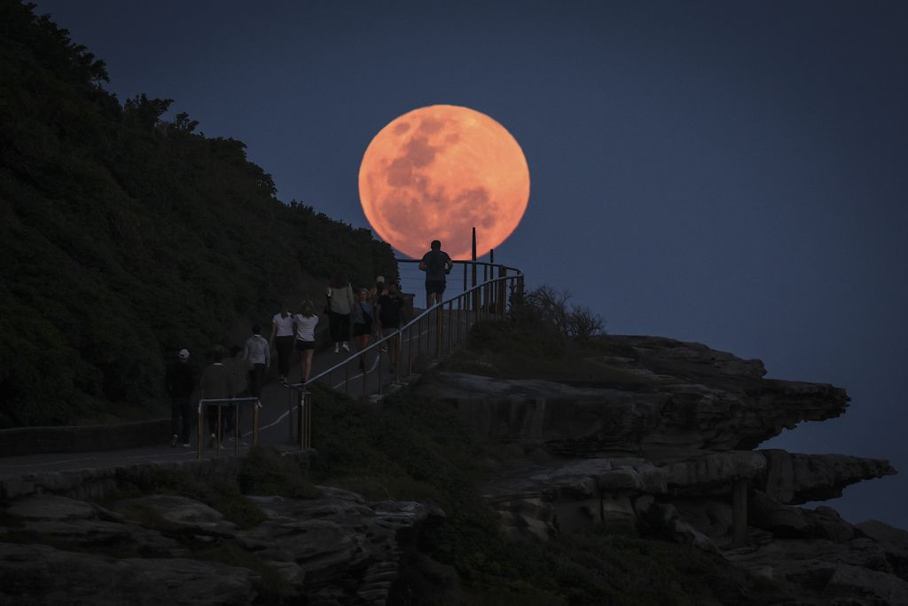 a supermoon rises and appears orange in front of a hillside near the coast, its light silhouetting a person climbing a staircase