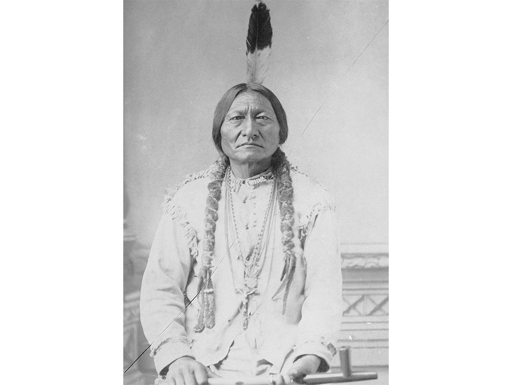 Historic black and white photograph of Sitting Bull sitting down with a feather in his hair and holding a pipe.