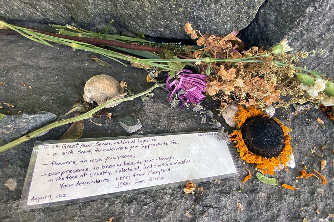 On a gray stone, a drying flower bouquet, a clam shell, and a laminated handwritten note: To 9th Great Aunt Sarah, victim of injustice: a silk scarf to celebrate your approach to life; flowers, to wish you peace; our presence, to bear witness to your stre