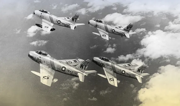 Four vintage fighter jets flying in a tight "finger-four" formation above a thick layer of white clouds in a black and white photograph.