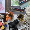 A variety of tools used to create art are displayed including scissors, fabric, a digital camera, tubes of paint and paintbrushes