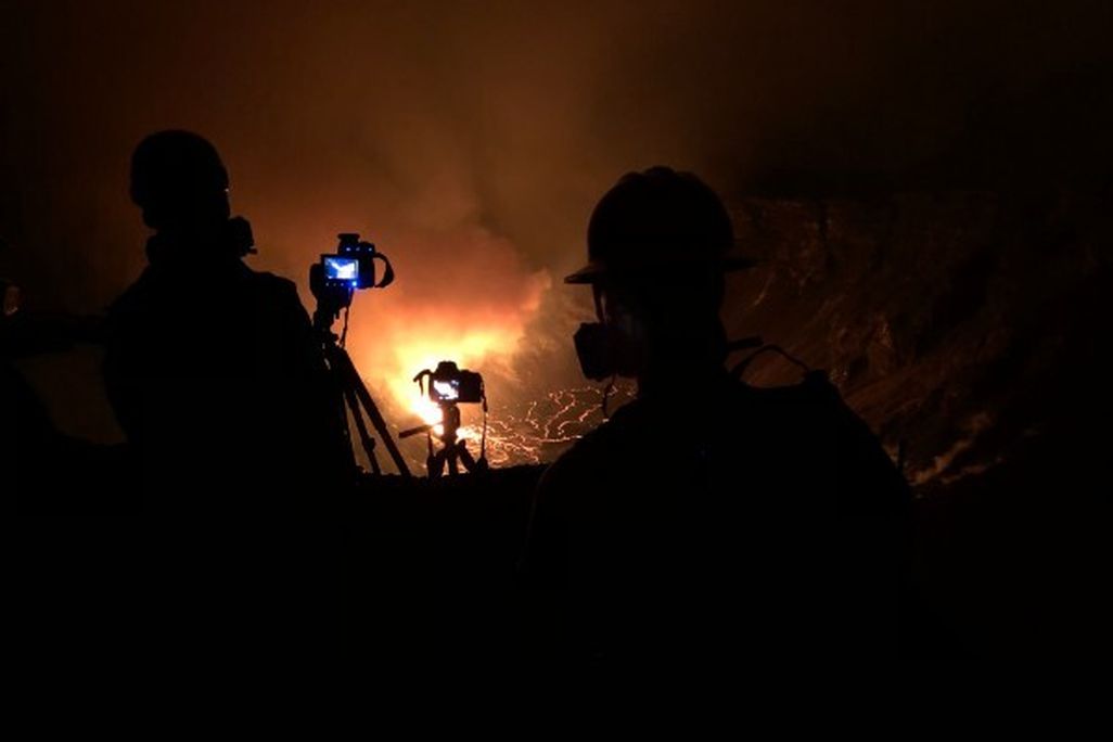 An image of the erupting volcano. Two people stand in the foreground with cameras, but only their silhouettes are visible. In the background, an orange cloud of steam is rising and the ground has bright orange crack in it. 