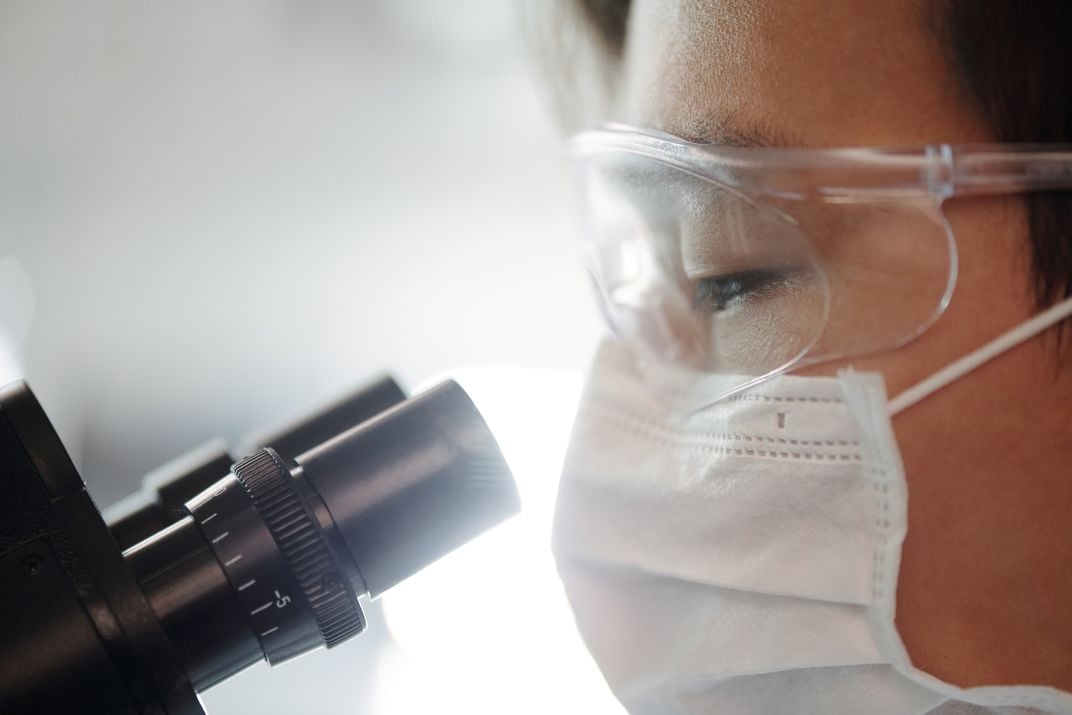 Scientist wearing protective mask and goggles looking at microscope