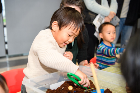A child smiles as he leans over a plastic tub filled with dirt using a set of tweezers to move objects around in the dirt.