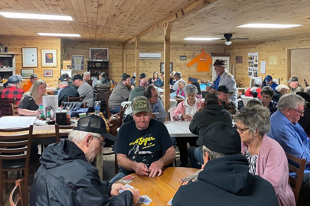 A few dozen people sit around tables playing cards in a wood-paneled room.