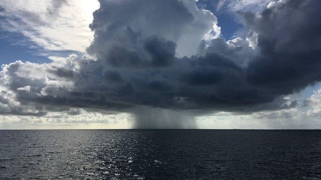 Rain falls from a small group of clouds over the open ocean as the sun shines behind the clouds.