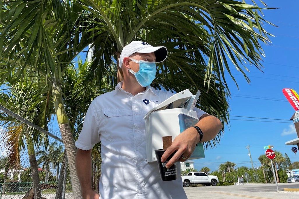 A man wearing a blue mask and a white baseball cap stands in front of a palm tree and holds a hexagon-shaped, toaster-sized box and a black cup