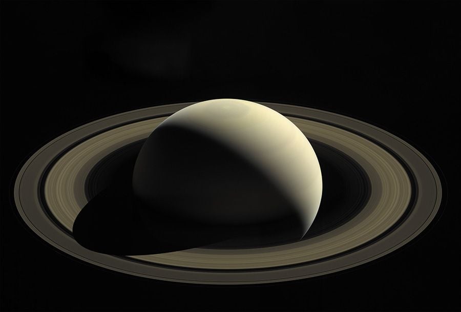 The smooth surface of a round planet is partially illuminated by the sun; the planet's other half is in shadow. The planet is surrounded by thousands of rings.