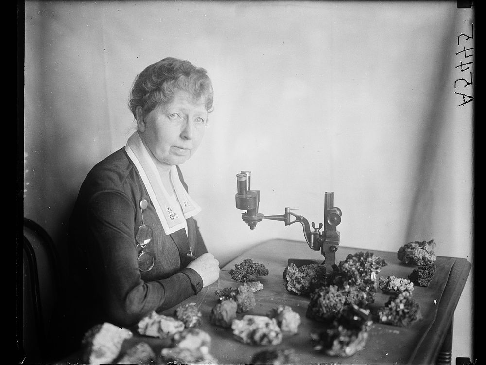 Black and white photo of Margaret W. Moodey sitting at a desk with a microscope and gemstones