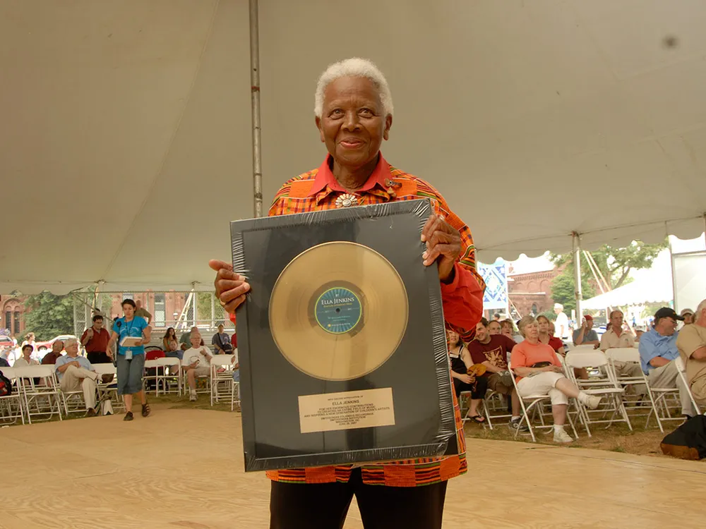 alt="An elder woman with dark skin and short white hair holds up a framed twelve-inch gold record, smiling. Behind her is a seated crowd under a festival tent."