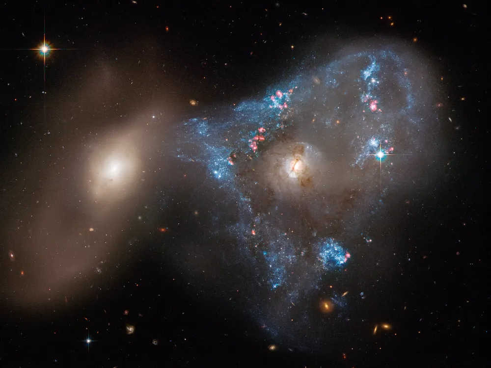 An image taken by the Hubble Space Telescope of two galaxies interacting with eachother. The galaxy on the right is surrounded by sparkling blue young stars. The galaxy towards the left is smaller and is not as flashy.