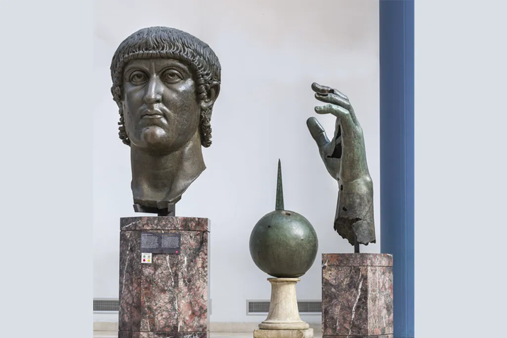 A view of a huge bronze cast head of a man with tightly curled hair and a serious expression, a sphere and his hand, all fragments that once were part of a huge 12m-tall sculpture
