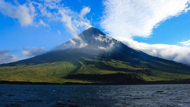 Cleveland Volcano with white clouds circling it as seen from the water on a bright, sunny day.