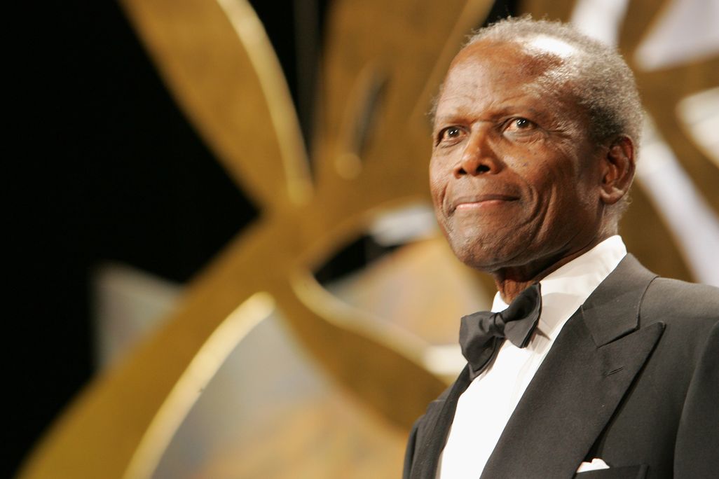 An elderly Poitier wears a black suit, white shirt and black bowtie and stands in front of a gold and silver background