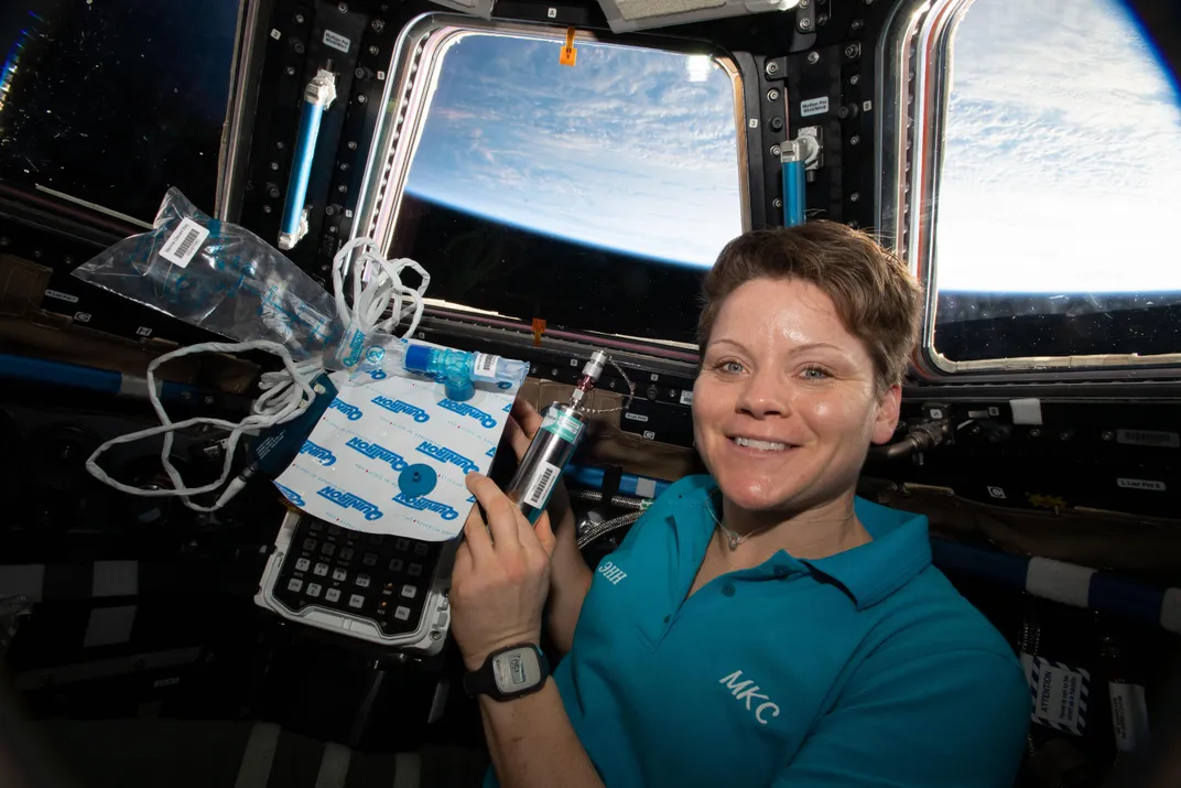 Female Astronaut Anne McClain holding biomedical gear for study while on the International Space Station.