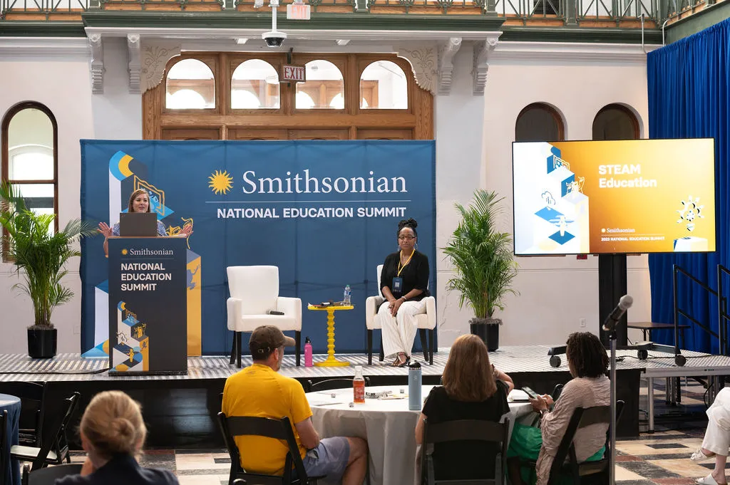 Two speakers on stage with a large banner behind them saying "Smithsonian National Education Summit"