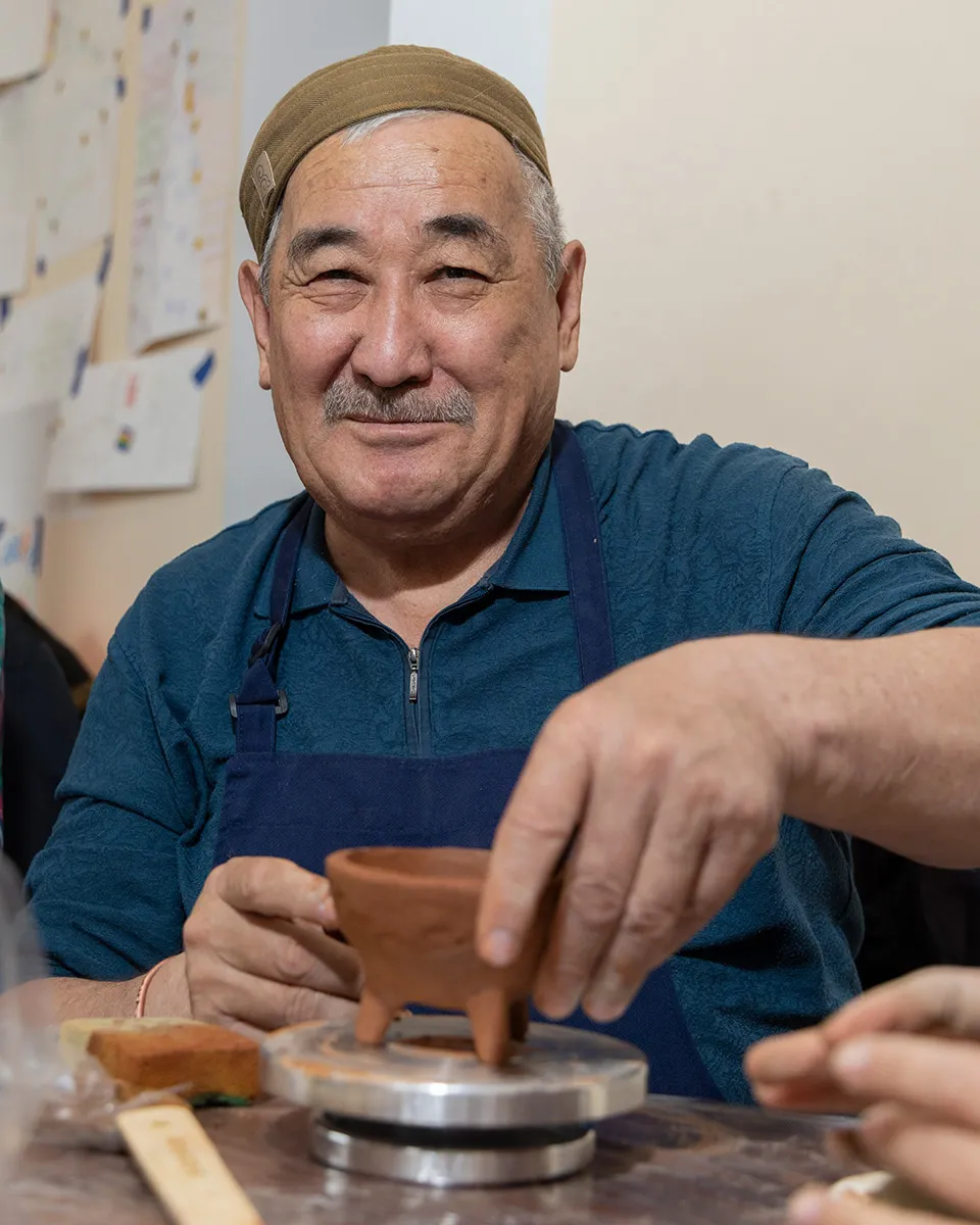 A man wearing a blue apron and beige cap smiles, holding a three-legged red clay pot.