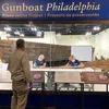 Visitors look into an exhibit window and see staff working on the wooden Gunboat