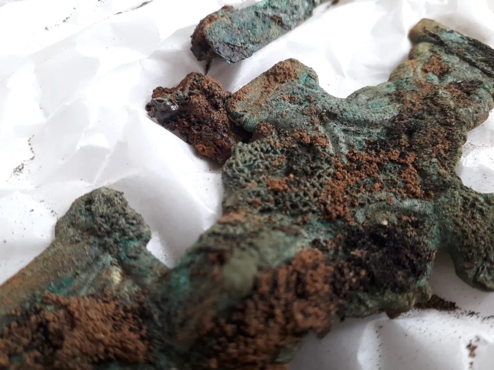 A close-up image of a greenish cross that appears carved and has bits of brown hardened material, textiles, adhering to its surface