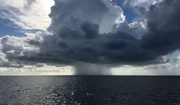 Rain falls from a small group of clouds over the open ocean as the sun shines behind the clouds.