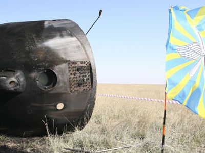 The Soyuz TMA-11 capsule lies on its side on the Kazakh steppe after landing. Search teams and companies involved in the rescue have planted their flags nearby.