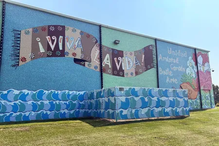 Turquoise mural covering the entire side of a building, with a pattern of waves along the bottom, and a brown textile with the Spanish words Viva la Vida! in the center.