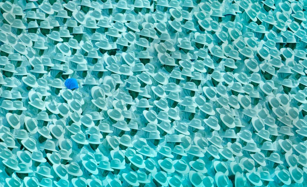 In a vibrant scene at Malaysia's National Independence Parade, hundreds of audience members are gathered, creating a unified sea of white as they wear matching hats and shirts in celebration. The bright white outfits reflect a sense of national pride and solidarity, contrasting against the colorful parade procession in the background. Among them, one person stands out—a lone figure without a hat, subtly breaking the uniformity. This small difference adds an intriguing detail to the image, drawing the viewer's eye amidst the cohesive crowd in this patriotic gathering.