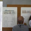 Backshot of two men work together to pin blueprints of a building design to a corkboard wall.