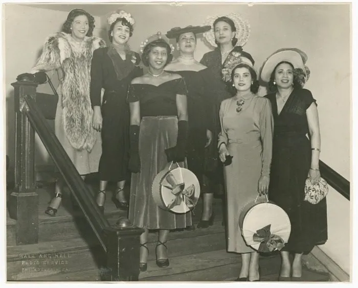 A black-and-white photograph of seven women standing on a flight of stairs. Mae Reeves stands to the farthest right on the lowest step and wears a white hat and floral patterned handbag.