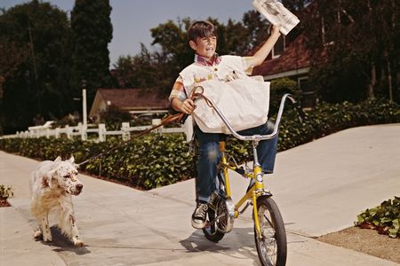 A boy riding his bike while delivering newspapers with his dog in tow, 1970s